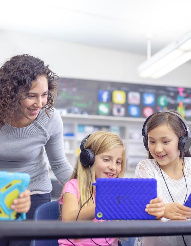 Profesora atendiendo a sus alumnas cómo ven contenidos digitales en sus tablets a las que están conectadas con cascos