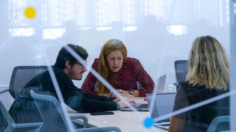 Tres personas trabajando con portátiles, dos de ellas consultando información en un ordenador