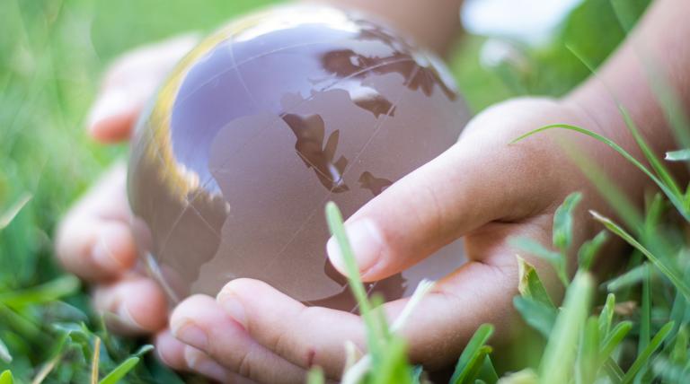 Manos sujetando una bola del planeta tierra traslucida, sobre un césped 